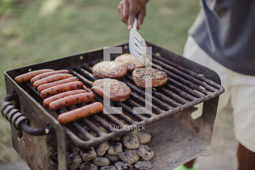 A man cooking hamburgers and hot dogs over coals on an outdoor barbecue grill.