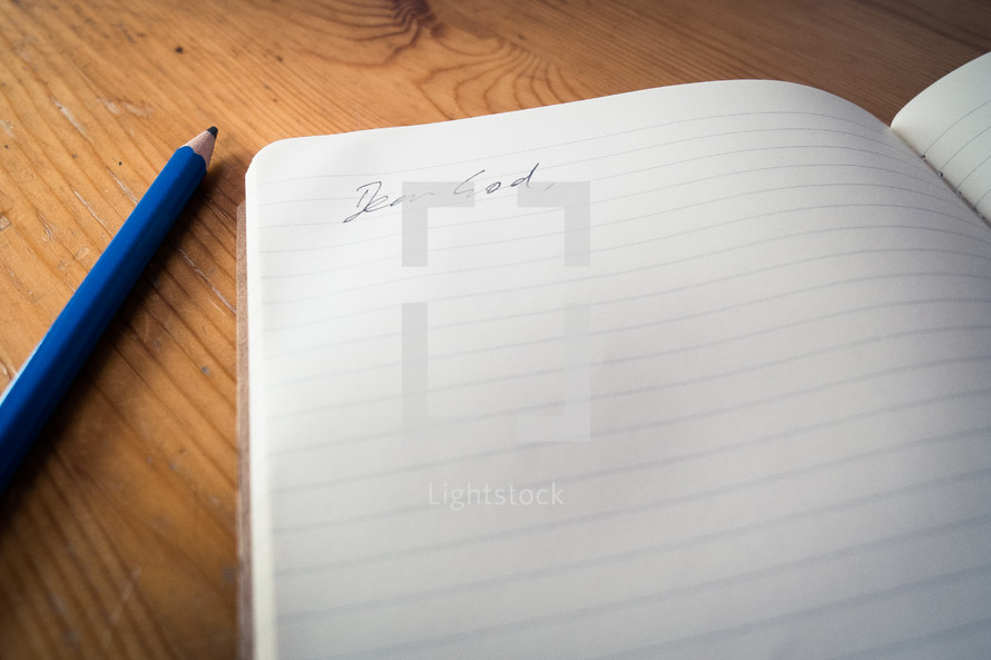 An open notebook with the words 'dear God' written, laying next to a blue pencil on a wooden desk