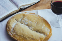 A loaf of bread and glass of wine on a table with an open bible