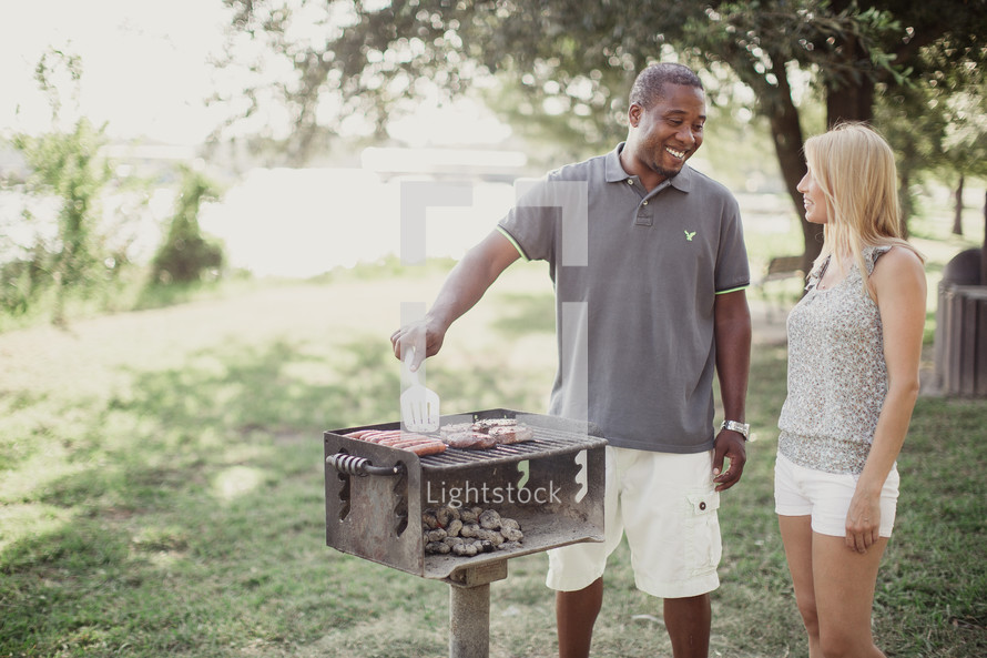A man talking to a woman while grilling hamburgers and hot dogs.