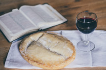 A loaf of bread, open bible, and a glass of wine on a table