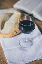 A glass of wine sits on a table next to a loaf of bread and an open bible.