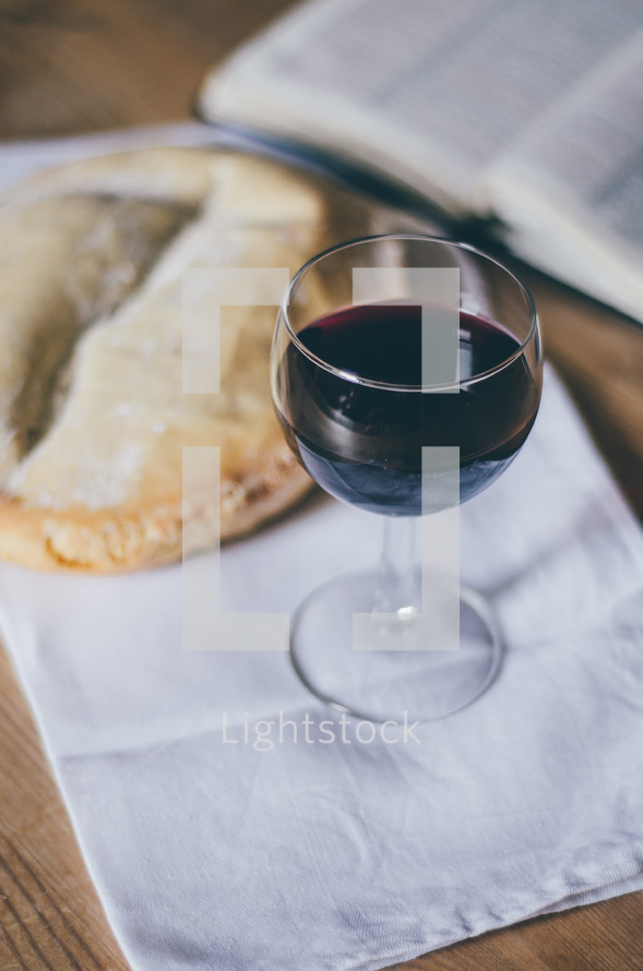A glass of wine sits on a table next to a loaf of bread and an open bible.