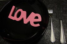 Pink letters spelling "love" on a black plate at a place setting.