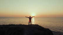 Silhouette of young successful traveler man standing on hill with open arms over sea at sunset.