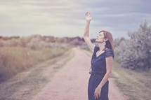 woman standing on a dirt road with her hand raised praising God 
