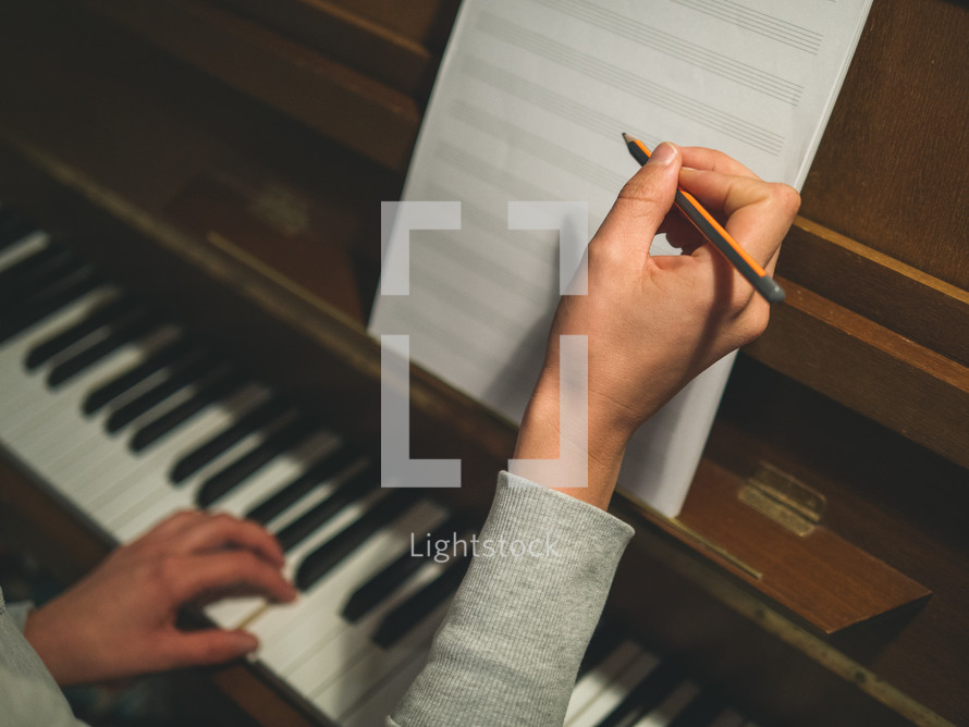 A woman's hands with pencil about to write on music notation paper, while playing a chord on the piano keyboard