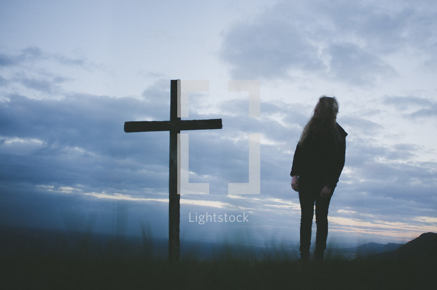 Silhouette of a persona standing by a cross in a field at sunrise.