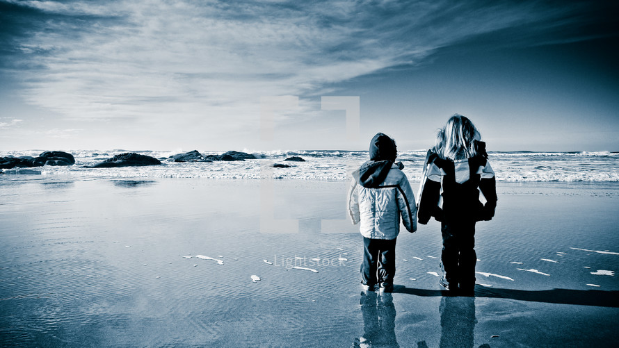 two children in winter coats standing on a beach