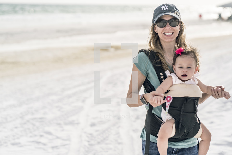 a mother walking on a beach with a baby in a baby carrier 
