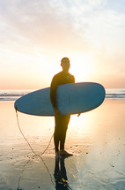 man in a wetsuit holding a surfboard on a beach at sunset 