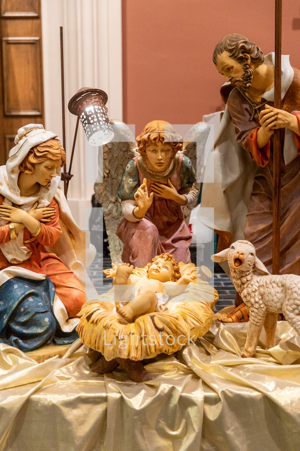 Statues of Mary, Joseph, and an angel facing baby Jesus in the manger; a little lamb stands on the side