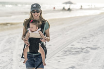 a woman walking on a beach with a baby in a baby carrier 