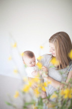 Mother holding infant daughter behind yellow flowers.