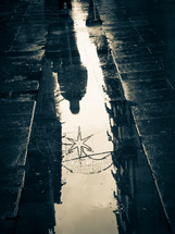 Duo-tone photo of man & star decoration in reflection in a puddle on a wet cobble-stone street.