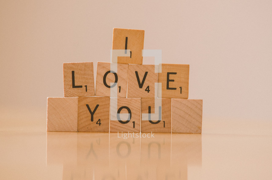 "I love you" arranged in stacked scrabble tiles on reflective table top.