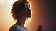 A woman woman prays with her hands folded and a light in back of her