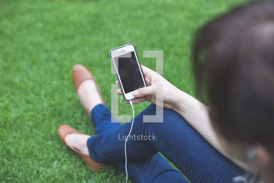 A girl sitting in grass and looking at a cell phone.