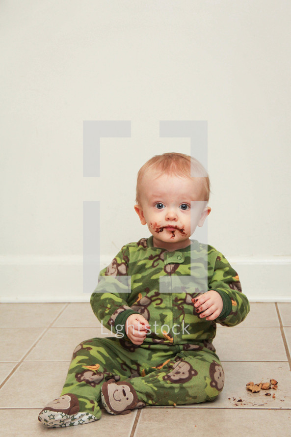 Toddler boy sitting on the floor eating a cookie, with a chocolate mess on his face.