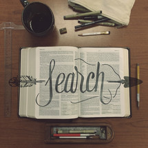 The word "search" over an open Bible on a table with writing utensils.