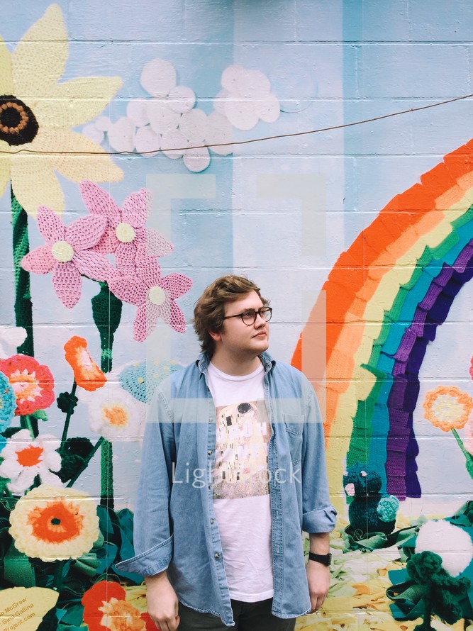a man standing in front of a artistically painted wall with rainbow and flowers 