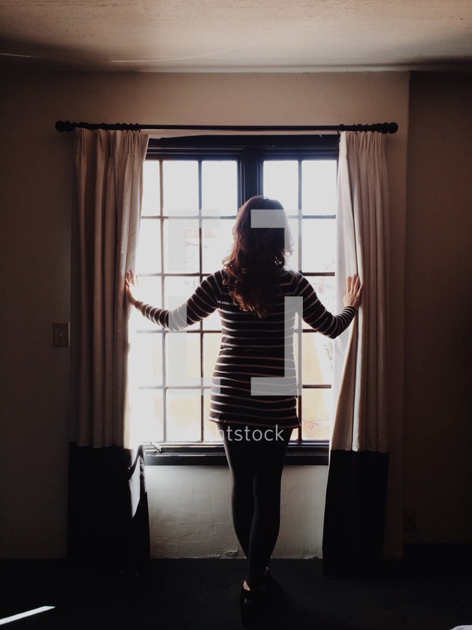 Woman opening the drapes and standing in front of the window.