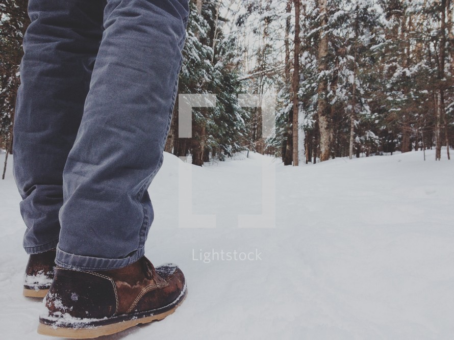A man's legs and feet standing in a snow covered forest.