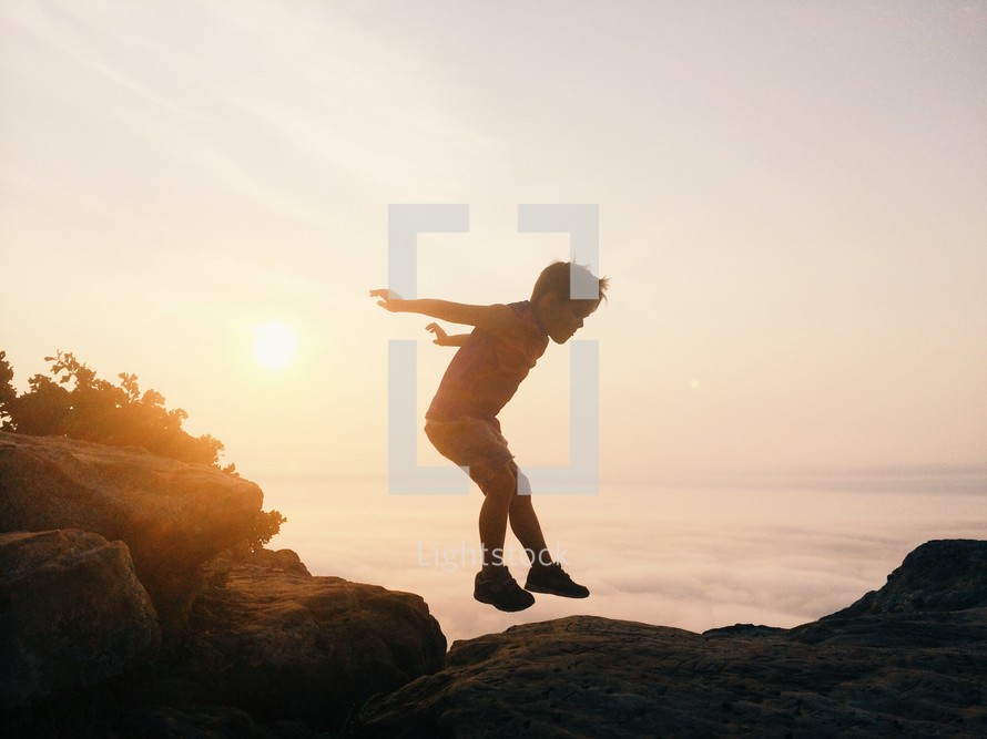 Silhouette of a boy jumping across rocks near the ocean at sunrise.