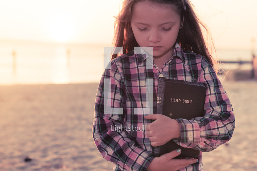 young girl thinking, standing on the beach holding her bible at sunset