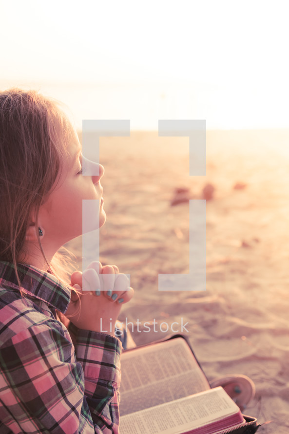young girl praying with bible in lap, sitting in the sand at the beach beach, sunset
