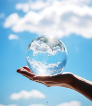Close up of human hand holding glass globe with blue sky background.