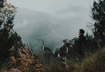 a man looking out at fog in a valley near a mountainside cliff 