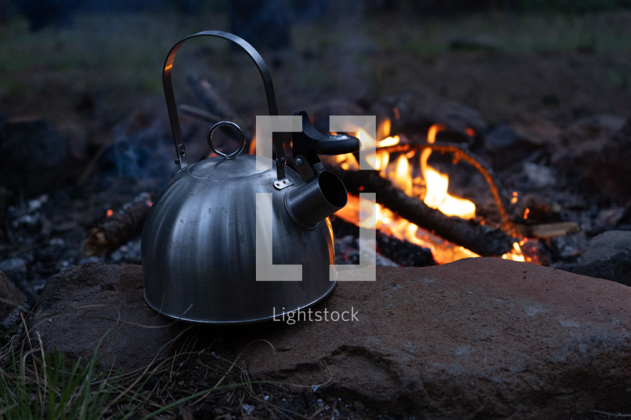 A tea kettle next to a camp fire at dusk