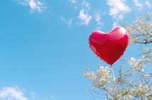 A heart-shaped balloon floating in a blue sky in a park during the day, creating a whimsical and romantic scene