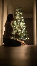 A woman kneeling in prayer in front of a Christmas tree 