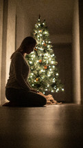 A woman kneeling in prayer in front of a Christmas tree 