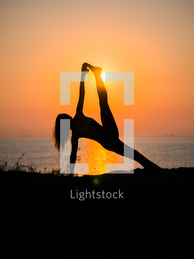Silhouette of young woman in swimsuit practicing yoga at amazing sunrise. Fitness, sport, yoga and healthy lifestyle concept. Girl doing a bridge on rock in sea.