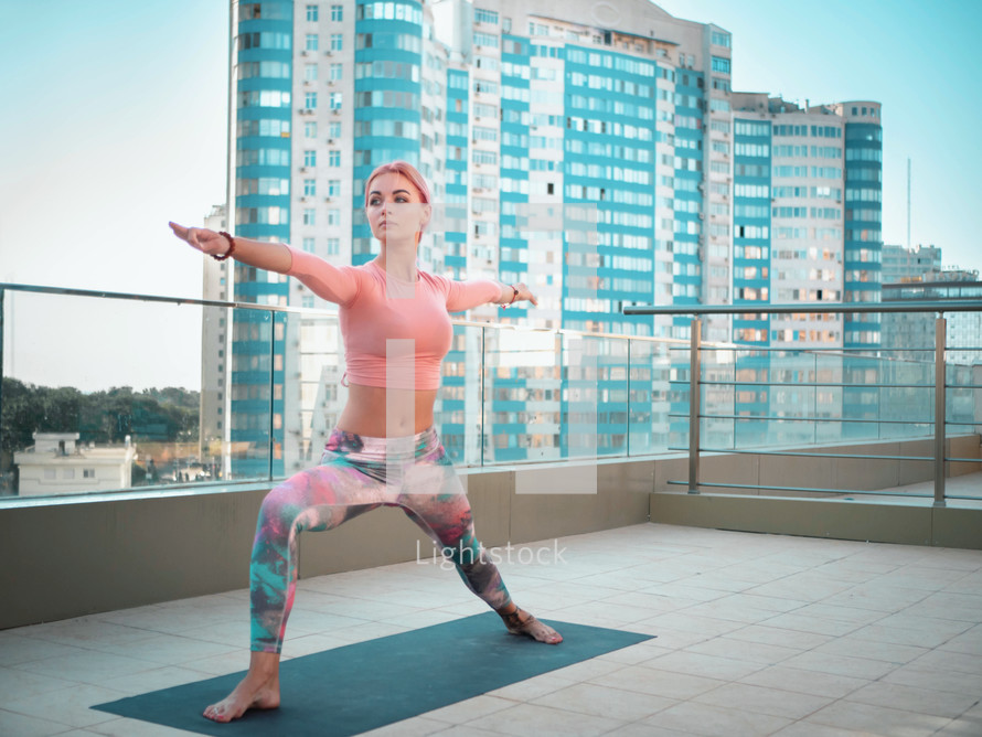  Young slim woman with pink dyed hair doing yoga practice on terrace of modern city. Girl keeping fit and healthy body relaxing on rooftop during practice pose