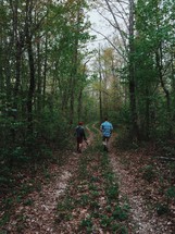 Two men walk along an overgrown road in the woods.