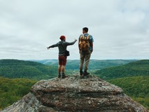 Two hikers standing on a rock pinnacle overlooking forest covered mountains.