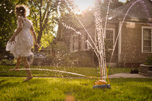 A little girl dances in the sprinklers in the warm golden light of summer.