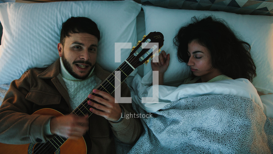 Man singing and playing the guitar in a bed while a woman sleeps