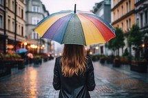 Girl walking in the rain through a gray city while carrying a rainbow umbrella. View from behind