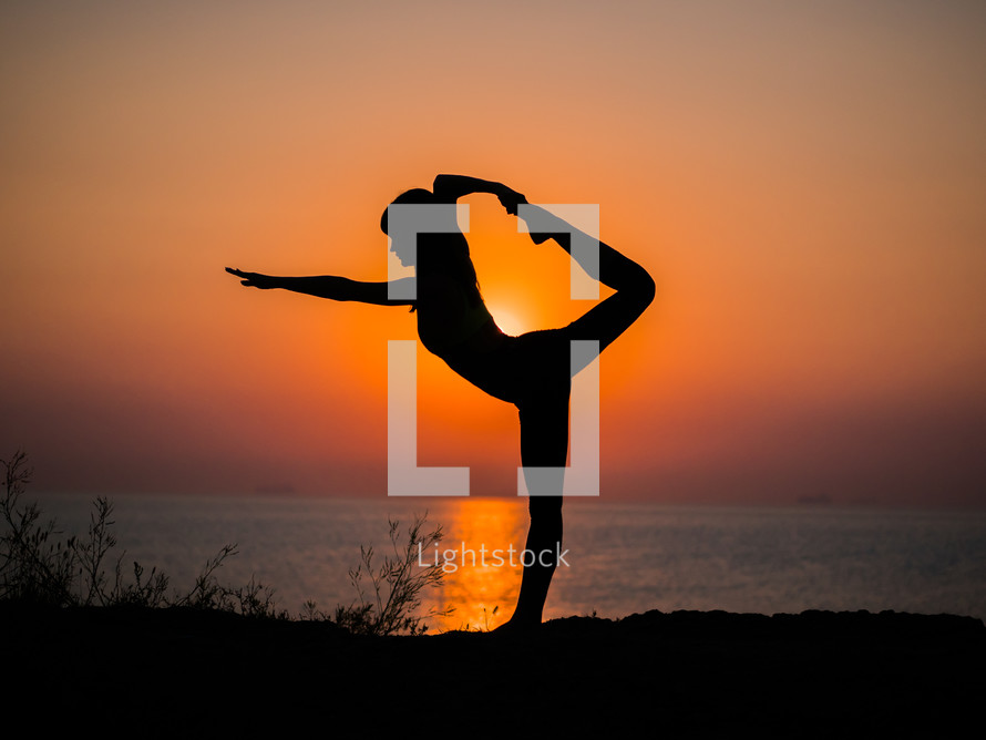 Silhouette of young woman in swimsuit practicing yoga at amazing sunrise. Fitness, sport, yoga and healthy lifestyle concept. Girl doing a bridge on rock in sea.