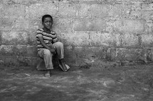 a shy little boy sitting on a rock in front of a wall outdoors 