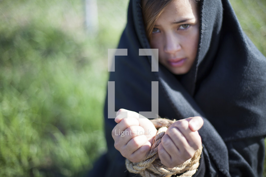 Girl in cape with hands bound with rope.