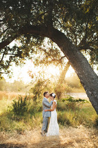 groom kissing his bride on the forehead standing under a tree outdoors