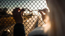 a woman holding reading glasses against a chain linked fence 