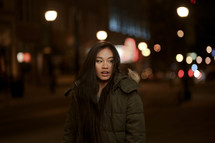 a young woman standing outdoors in a city at night in a coat 