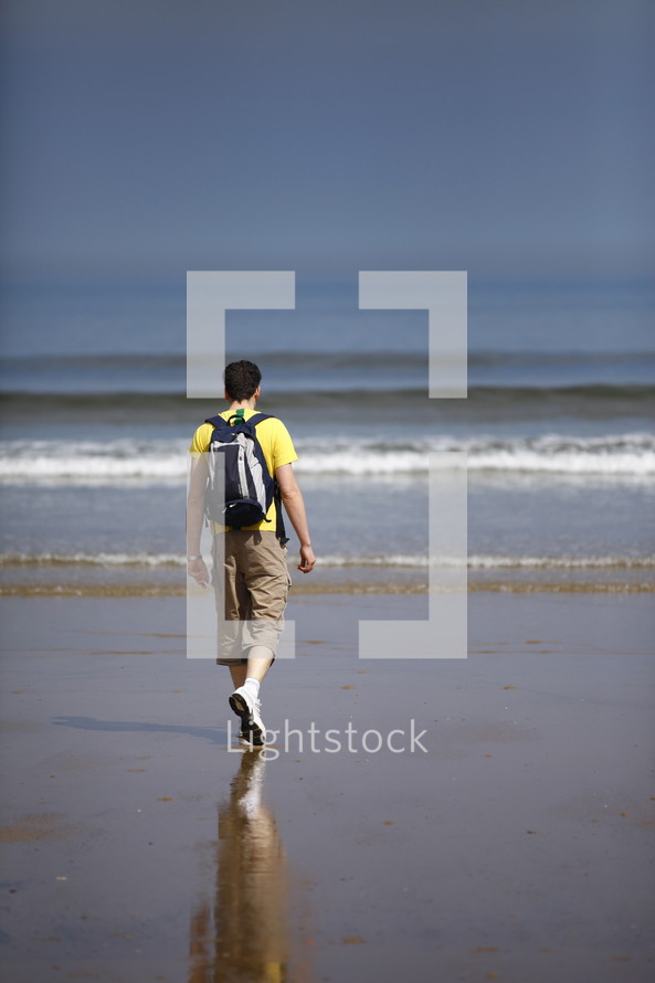 A man in a backpack walking along the beach.